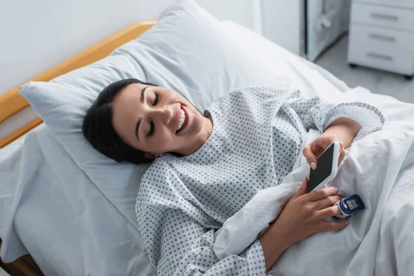 病院のベッドに横になっている間にスマートフォンを使って指に酸素濃度計をつけた陽気な女性 — ストック写真
