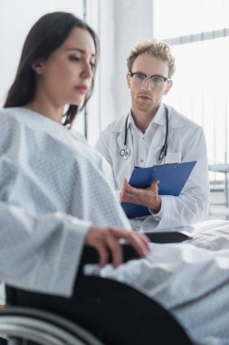 Kıvırcık doktor tekerlekli sandalyedeki engelli kadının yanında pano tutuyor. 