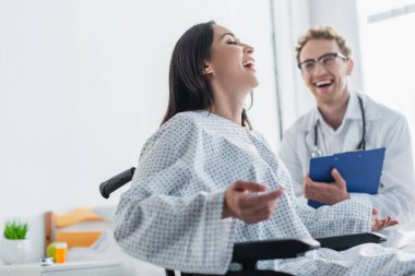 cheerful woman in wheelchair laughing near blurred doctor  clipart
