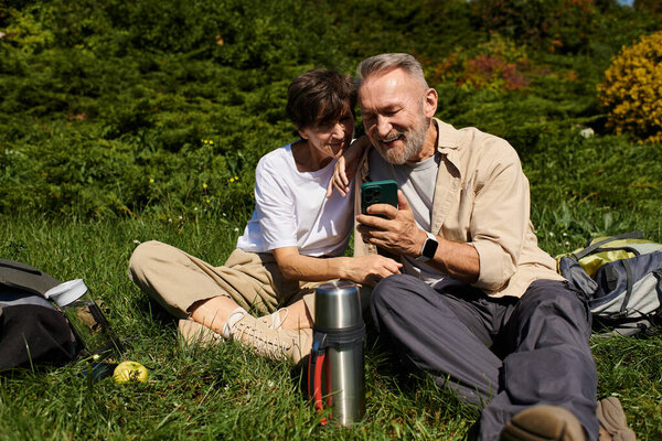 Старшая пара отдыхает в горах, делится улыбкой и проверяет телефон..