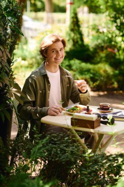 A young man with a cheerful expression eats a tasty meal in a lively cafe, surrounded by plants.