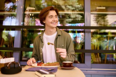 In a cozy modern cafe, a young man in an olive green shirt smiles as he enjoys his meal.