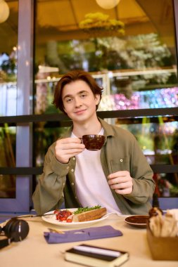 A young man in an olive green shirt sits in a modern cafe enjoying his meal and drink.