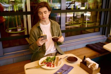 A young man in an olive green shirt relaxes with food and coffee in a modern cafe.