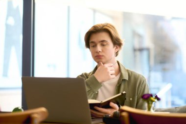 A young man sits in a trendy cafe, engaged with his laptop as he studies and takes notes.