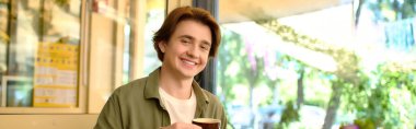 A young man in a casual shirt smiles in a lively cafe, banner