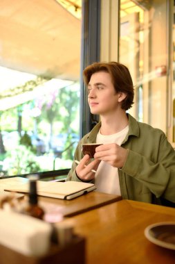 In a vibrant cafe, a young man sips coffee, deeply engaged in his thoughts.