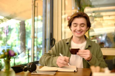 A young man in a shirt enjoys coffee and taking notes at a trendy cafe.