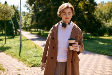 A young man in a trendy coat strolls through an autumn park, sipping coffee and enjoying the day.