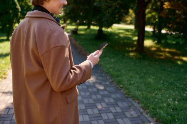 A young man in a trendy coat explores a vibrant autumn park while checking his phone.