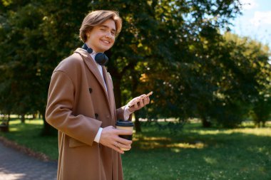 A young man in a fashionable coat strolls through an autumn park while sipping coffee.