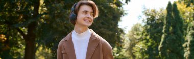 A young man strolls through a vibrant autumn park, dressed in a stylish coat and headphones, banner