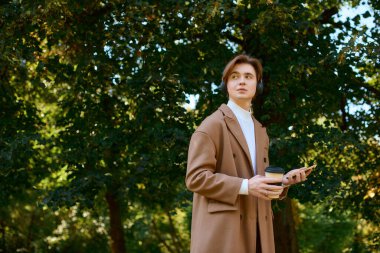 A young man in a chic coat relaxes outdoors surrounded by vibrant autumn leaves.