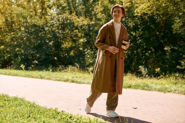 A young man dressed in a stylish coat walks leisurely in a vibrant autumn park.