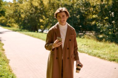 A young man in a fashionable coat walks through an autumn park, sipping a drink and using his phone.