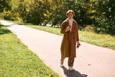 A young man in a fashionable coat strolls through a vibrant autumn park.