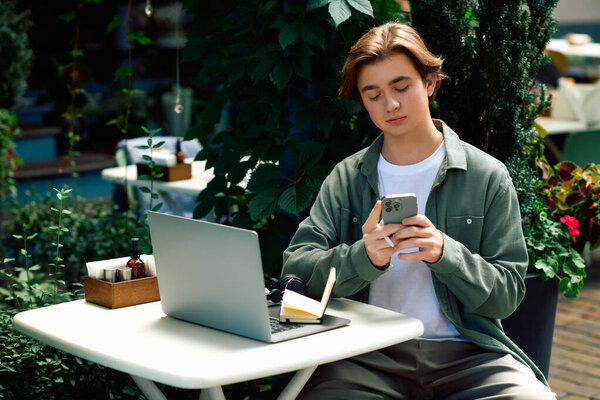In a chic cafe, a young man in a shirt works on his laptop and occasionally checks his phone.