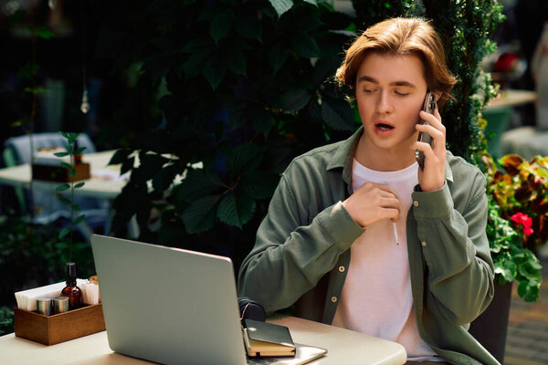 A focused young man in a casual shirt engages in a phone call while working on his laptop.