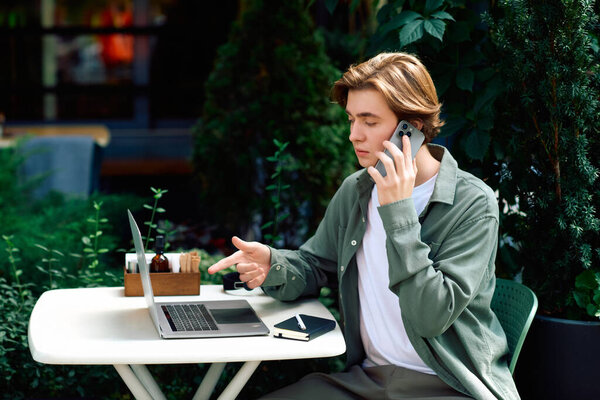 A young man in a modern cafe engages in online work while using his laptop and phone.