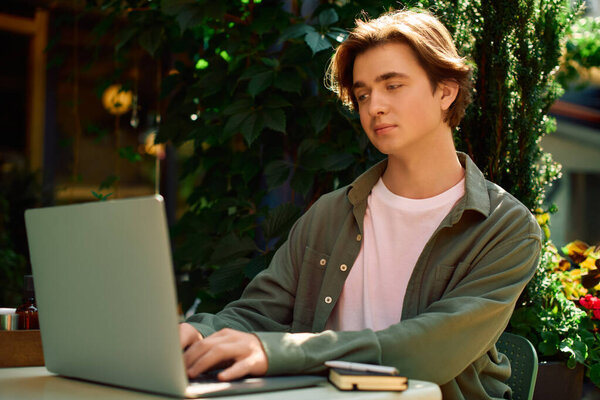 A young man in a shirt works on his laptop at a sunny cafe surrounded by greenery.