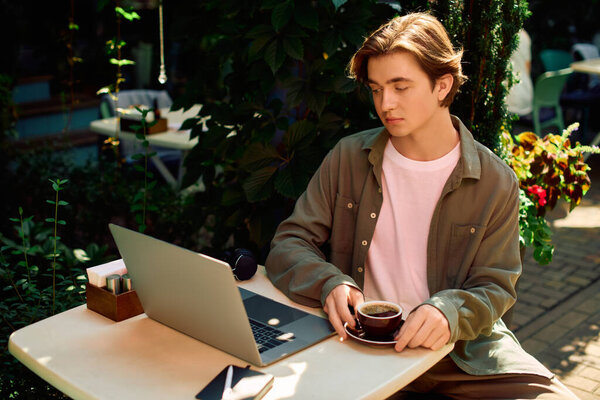 A young man in a shirt focuses on his laptop at a vibrant cafe, sipping coffee.