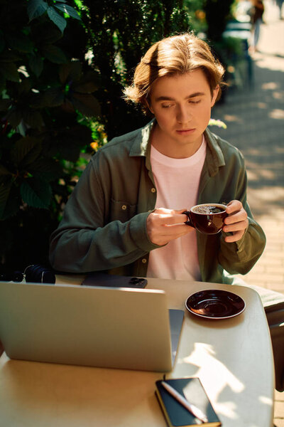 A young man in a shirt sits at a cafe, focused on his laptop while sipping a warm beverage.