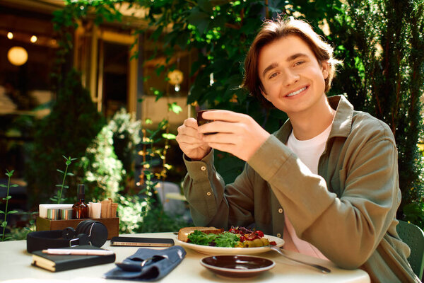 A young man in an olive green shirt smiles while enjoying his meal at a modern cafe outdoors.