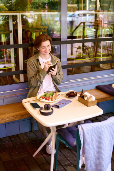 A young man in an olive green shirt engages with his phone while eating in a stylish cafe.