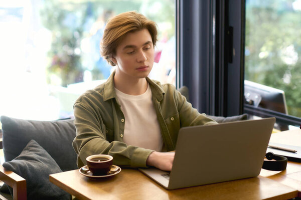 A young man in a casual shirt works on his laptop in a modern cafe, enjoying his coffee.