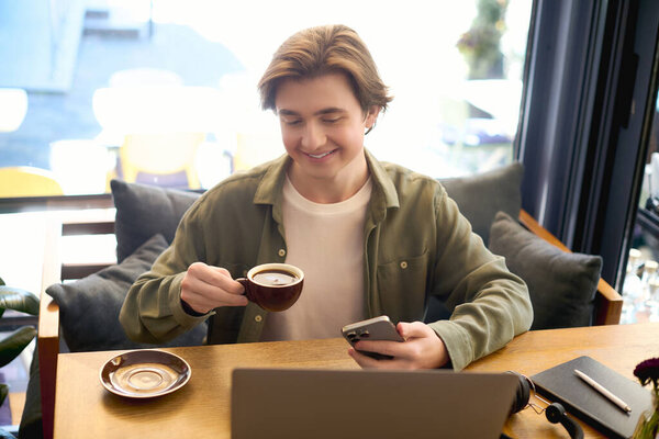 A young man in a shirt smiles as he works on his laptop while enjoying coffee in a lively cafe.