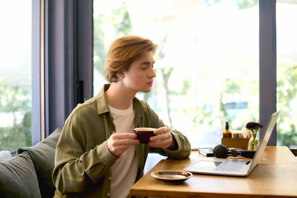 A young man in a shirt focuses on his laptop, savoring coffee in a stylish cafe.