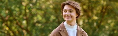 A young man dressed in a stylish coat smiles while relaxing in an autumn park, banner