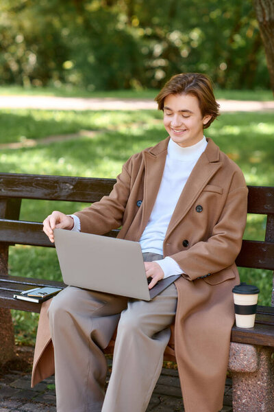 A young man in a chic coat works on his laptop, savoring the autumn vibes in a serene park.