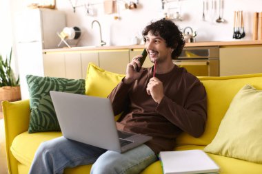 A handsome young man chats on the phone while using his laptop on a yellow sofa in a modern kitchen.