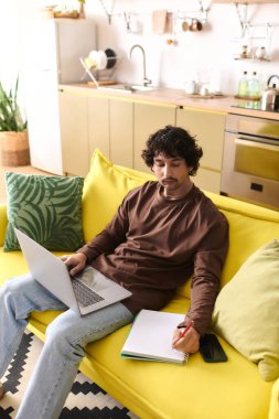 A young man on a cozy yellow couch writes notes on his laptop in a stylish kitchen.