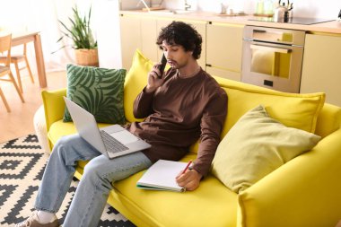 Handsome young man talks on the phone and takes notes on a cozy yellow sofa at home.
