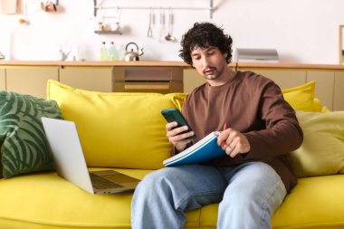 A young man sits on a bright yellow couch, focused on his smartphone while jotting notes.