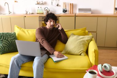 Young man engaged in a phone conversation while taking notes at home, surrounded by warm decor.