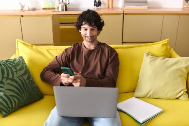 Handsome young man smiles while checking his smartphone and working on a laptop in a bright room.