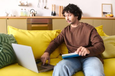 A young man engages in study at home, balancing a laptop and a book while seated on a bright couch.
