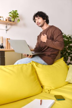 A young man sits on a bright yellow couch, focused on his laptop while deep in thought.
