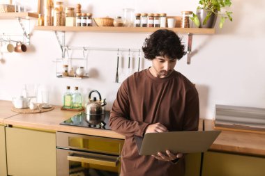 Handsome young man focuses on his laptop as he prepares a meal in a modern kitchen space.