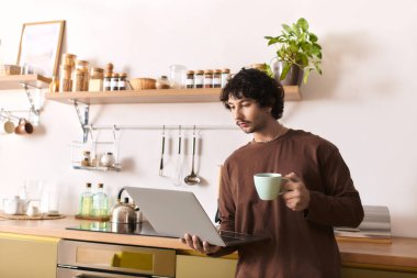Young man holds a cup, focused on his laptop in a warm, inviting kitchen space.