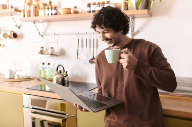 Handsome young man smiles while holding a cup and using his laptop in a cozy kitchen.