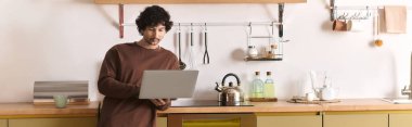 Young man with curly hair focuses on his laptop in a stylish kitchen setting.
