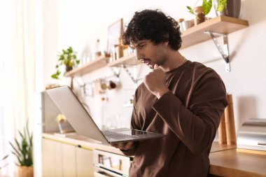 Handsome young man focuses on his laptop in a warm, inviting kitchen filled with plants.