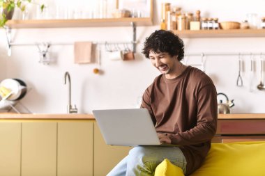 A handsome young man smiles as he engages with his laptop in a warm, inviting kitchen.
