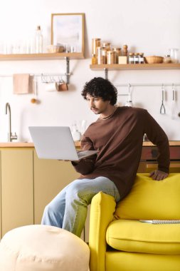 Handsome young man focuses on his laptop, seated on a bright yellow sofa in a modern kitchen.