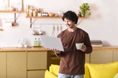 Cheerful young man with curly hair works on his laptop, sipping coffee in a cozy living space.