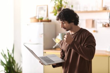 Young man in casual attire savors coffee as he focuses on his laptop in a bright kitchen.